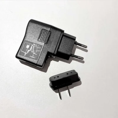 A detached black European plug adapter with two round pins, part of the ComPilot Power Supply, rests on a white surface. The main unit features a diagram demonstrating how to attach the plug section.
