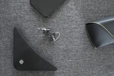 A smartphone, wireless earpiece, Widex TV Play device, triangular black speaker, and a black case with yellow stitching are displayed on a textured gray surface.