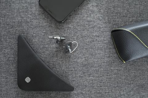 A smartphone, wireless earpiece, Widex TV Play device, triangular black speaker, and a black case with yellow stitching are displayed on a textured gray surface.
