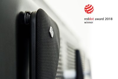 Close-up of a textured black speaker grille with a small logo near a wall. On the right, the Widex TV Play product name and Red Dot Award 2018 winner logos appear on a white background.