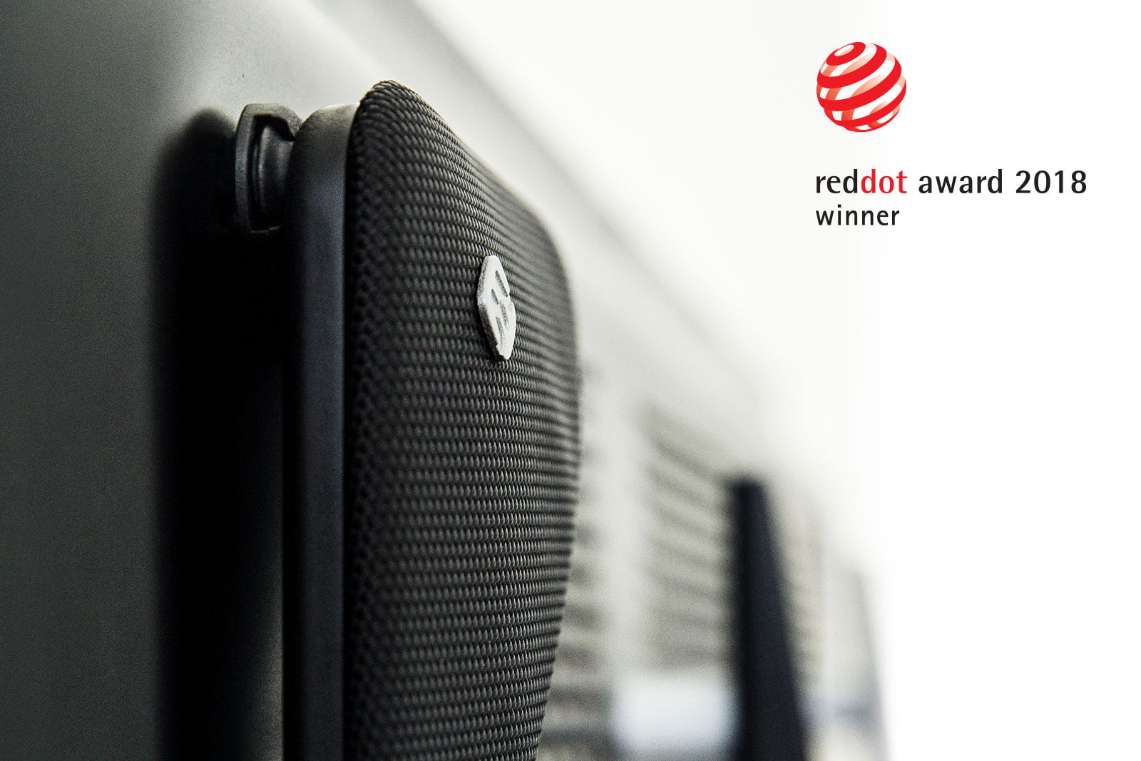 Close-up of a textured black speaker grille with a small logo near a wall. On the right, the Widex TV Play product name and Red Dot Award 2018 winner logos appear on a white background.